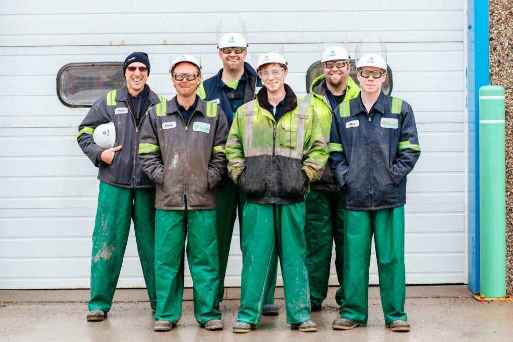Six industrial workers in safety gear smiling in front of a garage door.